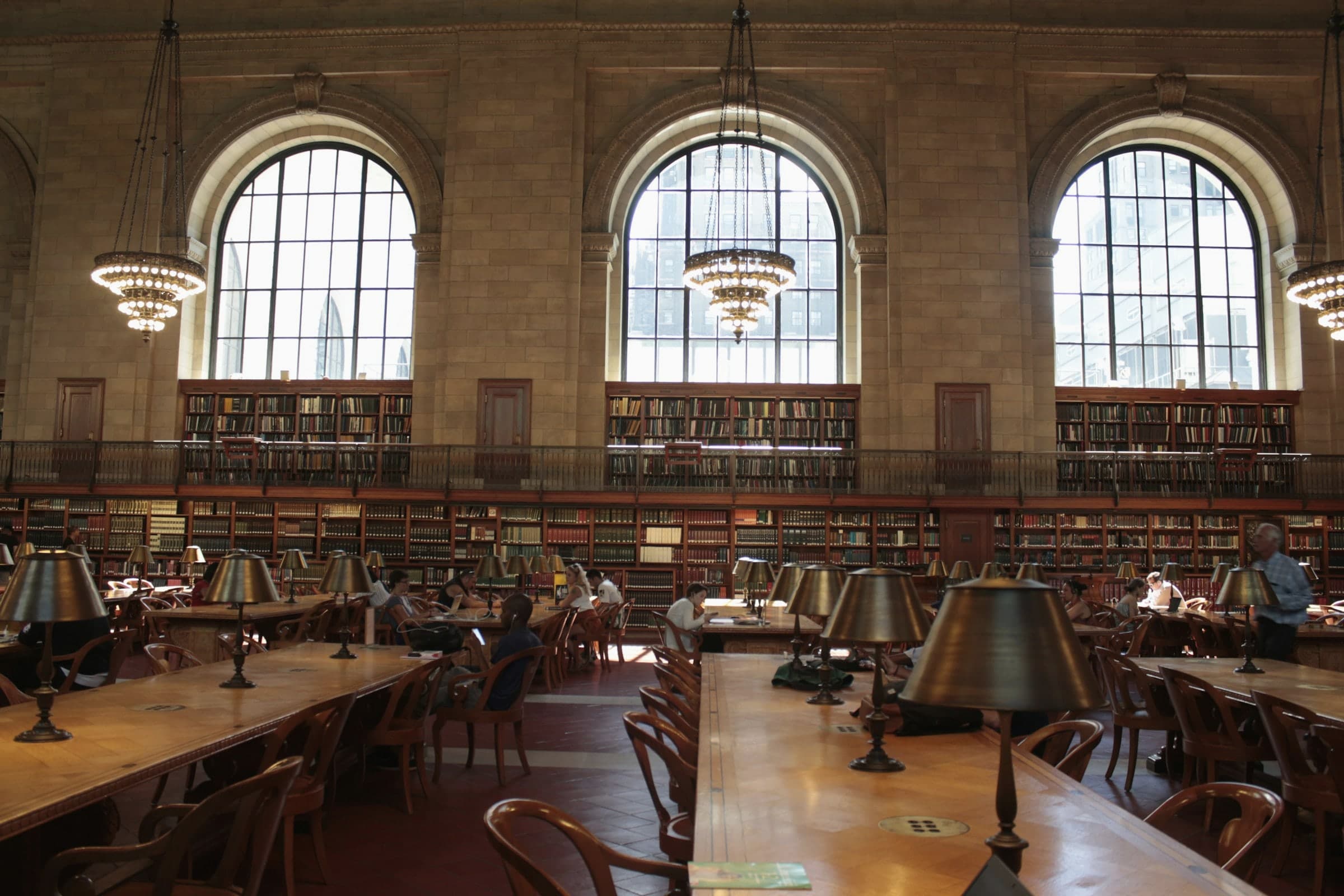 Großer Lesesaal einer Bibliothek mit Bogenfenstern, Bücherregalen, Messinglampen und lesenden Menschen an langen Tischen