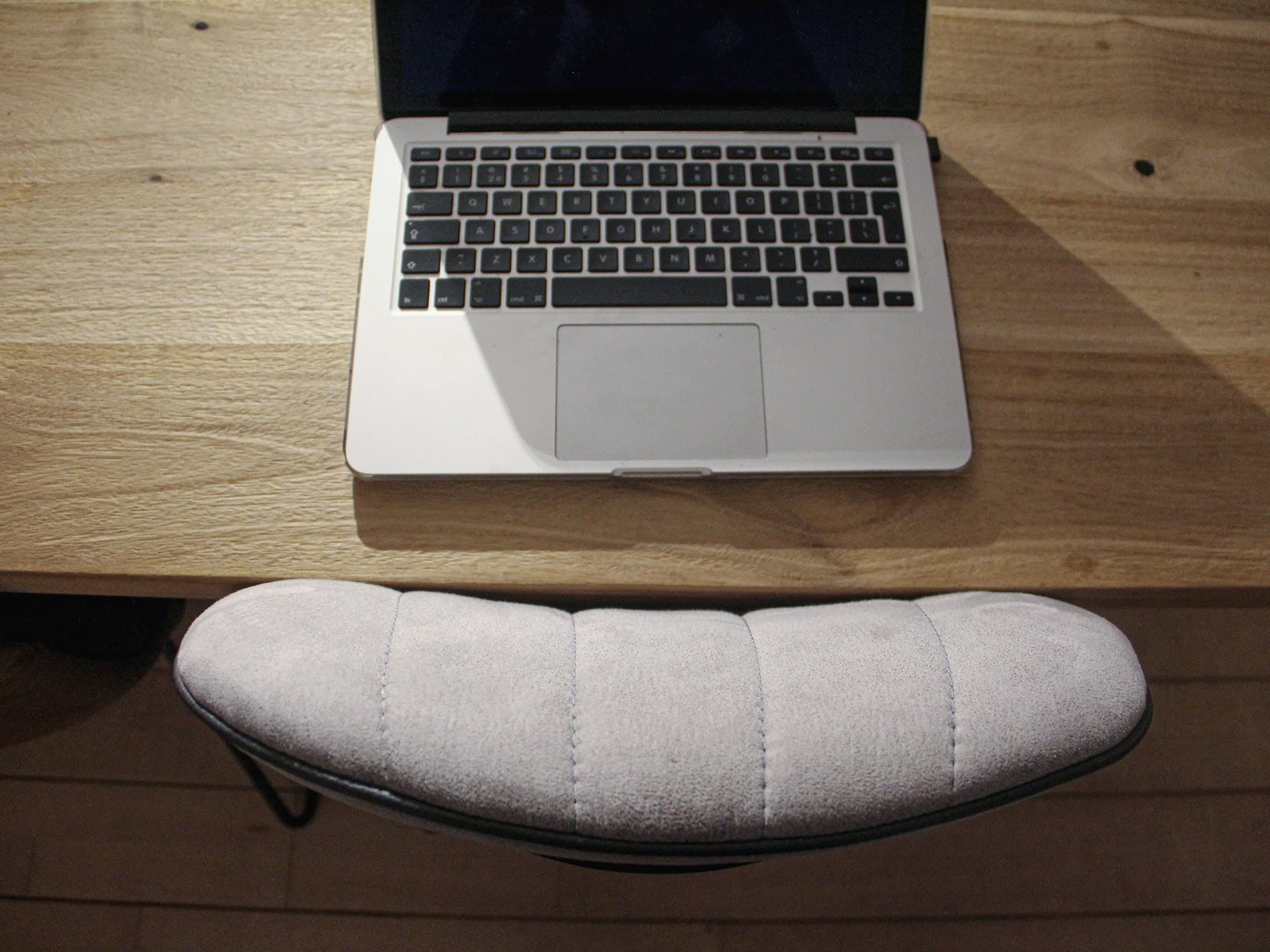 Aufgeklappter Laptop mit dunklem Bildschirm auf einem sonnigen Eichentisch, davor ein gepolsterter Stuhl, von oben gesehen