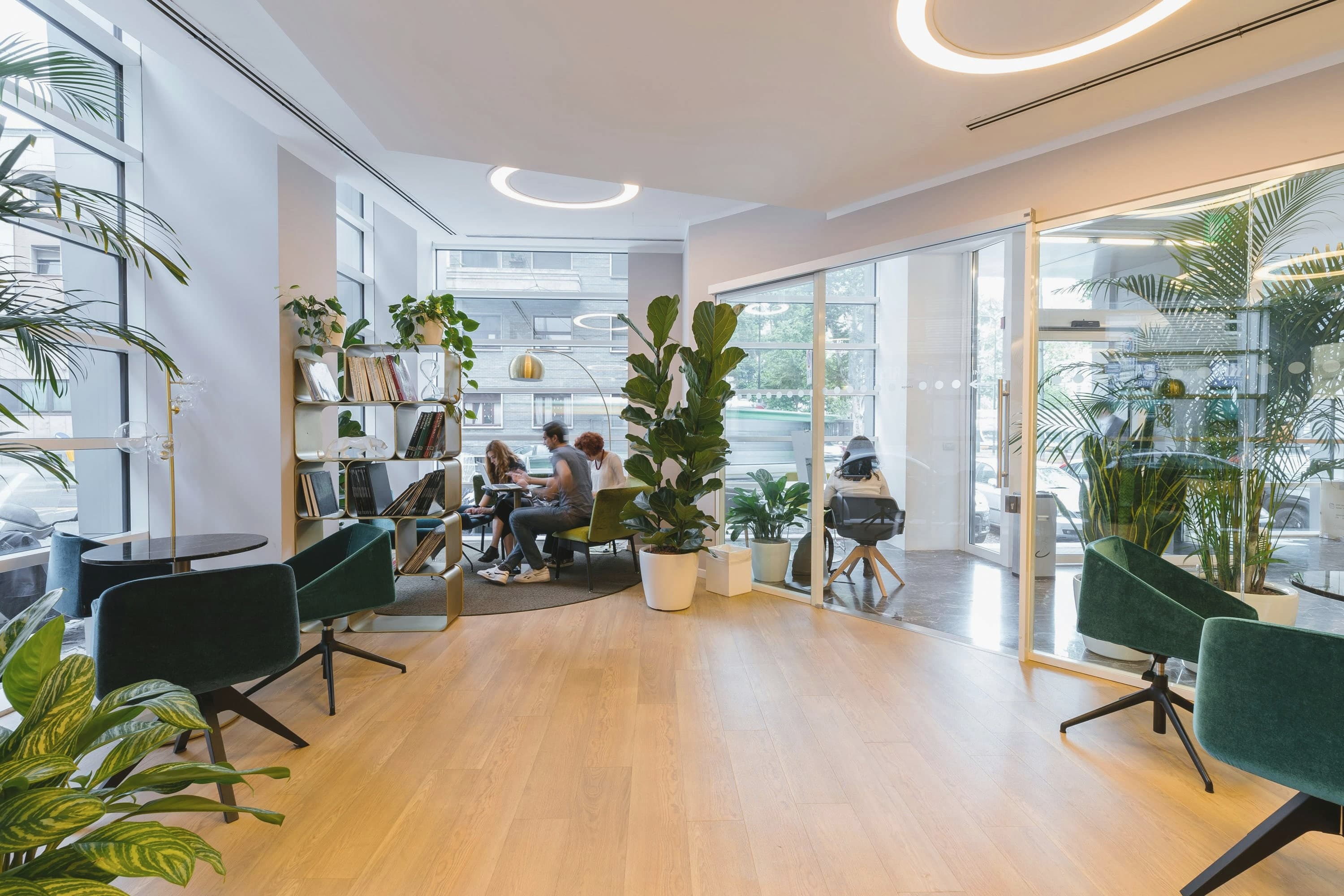 Offene Bürolounge mit Eichenparkett, grünen Samtsesseln, Pflanzen und einem Regal, im Hintergrund arbeitet ein kleines Team